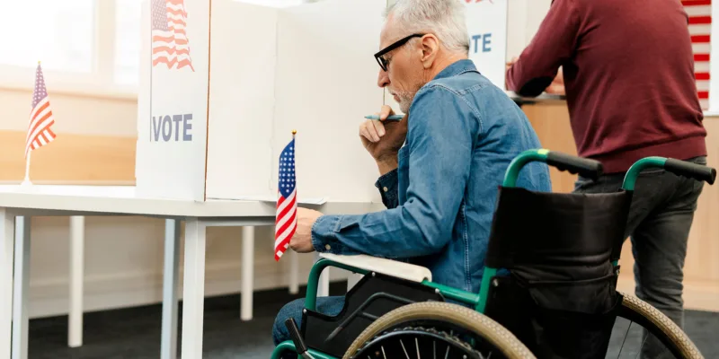 An older man in a wheelchair voting at a polling station, holding a pen and sitting at a booth with an American flag and the word "VOTE." Another person stands voting at a booth nearby.