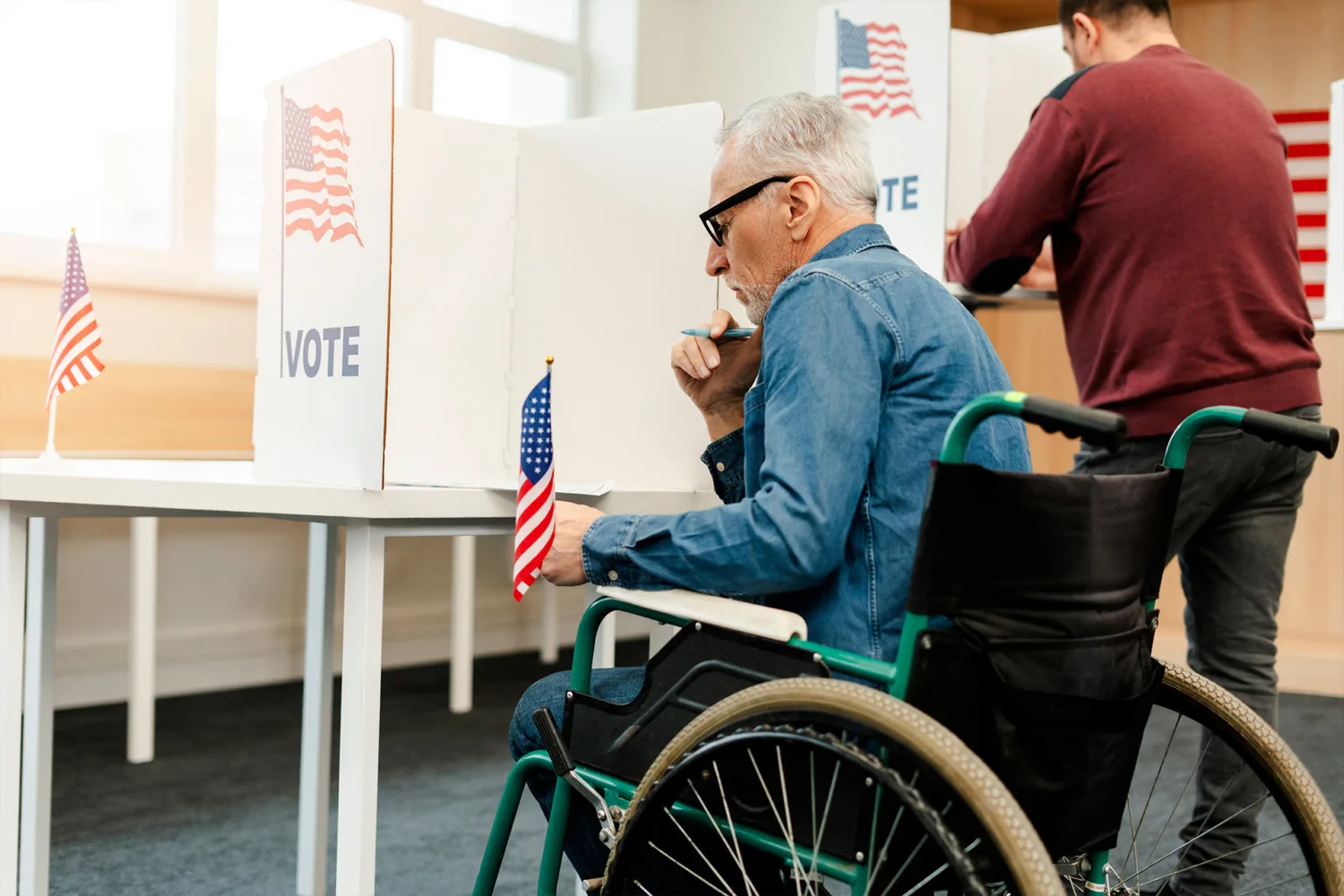 An older man in a wheelchair voting at a polling station, holding a pen and sitting at a booth with an American flag and the word "VOTE." Another person stands voting at a booth nearby.