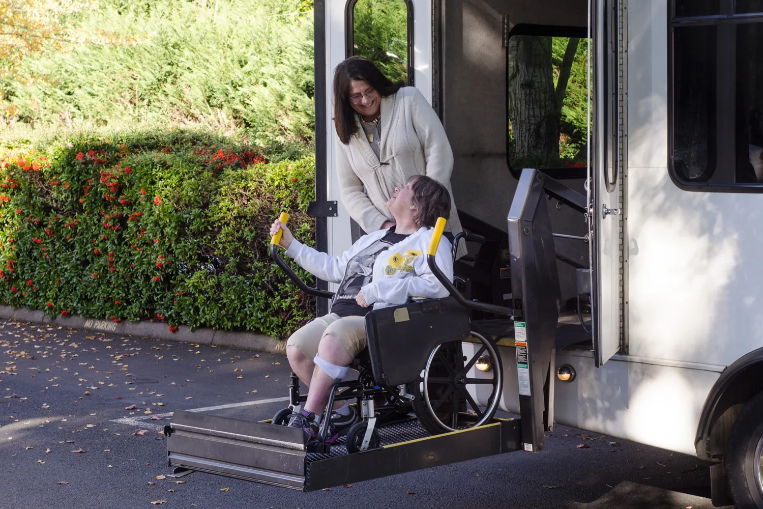 A woman in a wheelchair uses a lift to exit a van, smiling at another woman standing nearby. The scene is outdoors, with greenery and a sunny atmosphere.