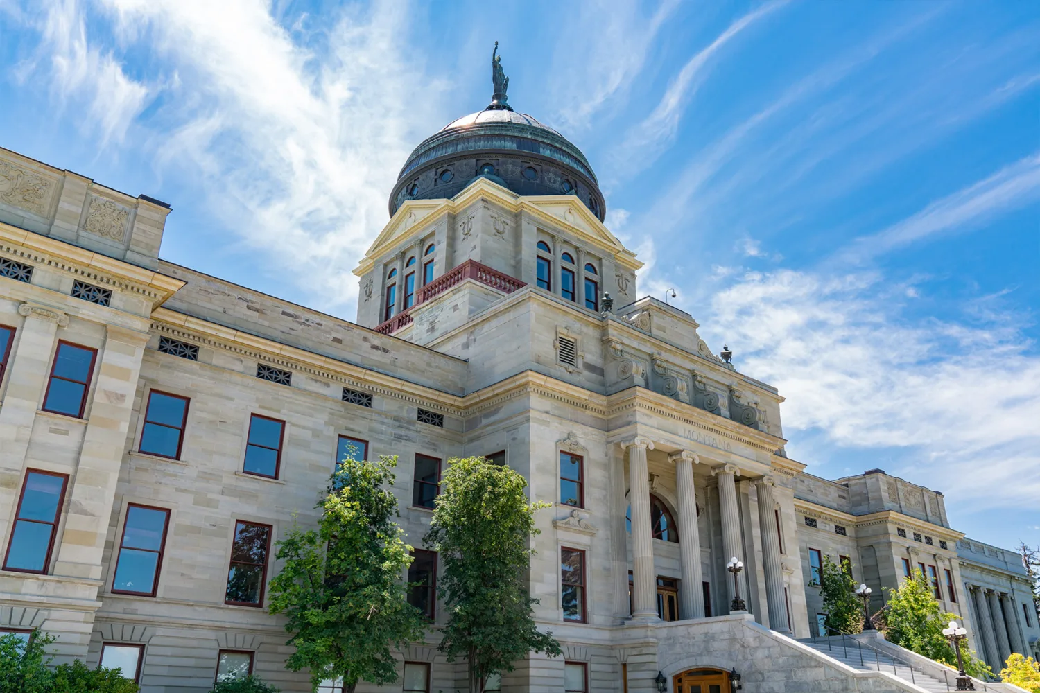 A grand stone building with columns and a large domed roof stands under a blue sky with wispy clouds; trees line the front, and a statue tops the dome.