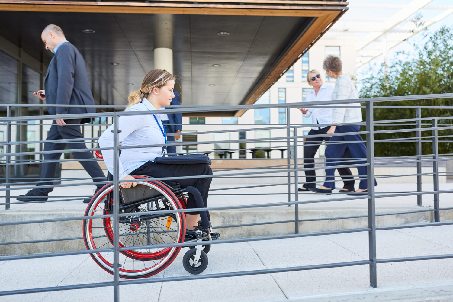 A woman in business attire uses a wheelchair on a ramp outside a modern building, while three people walk nearby.