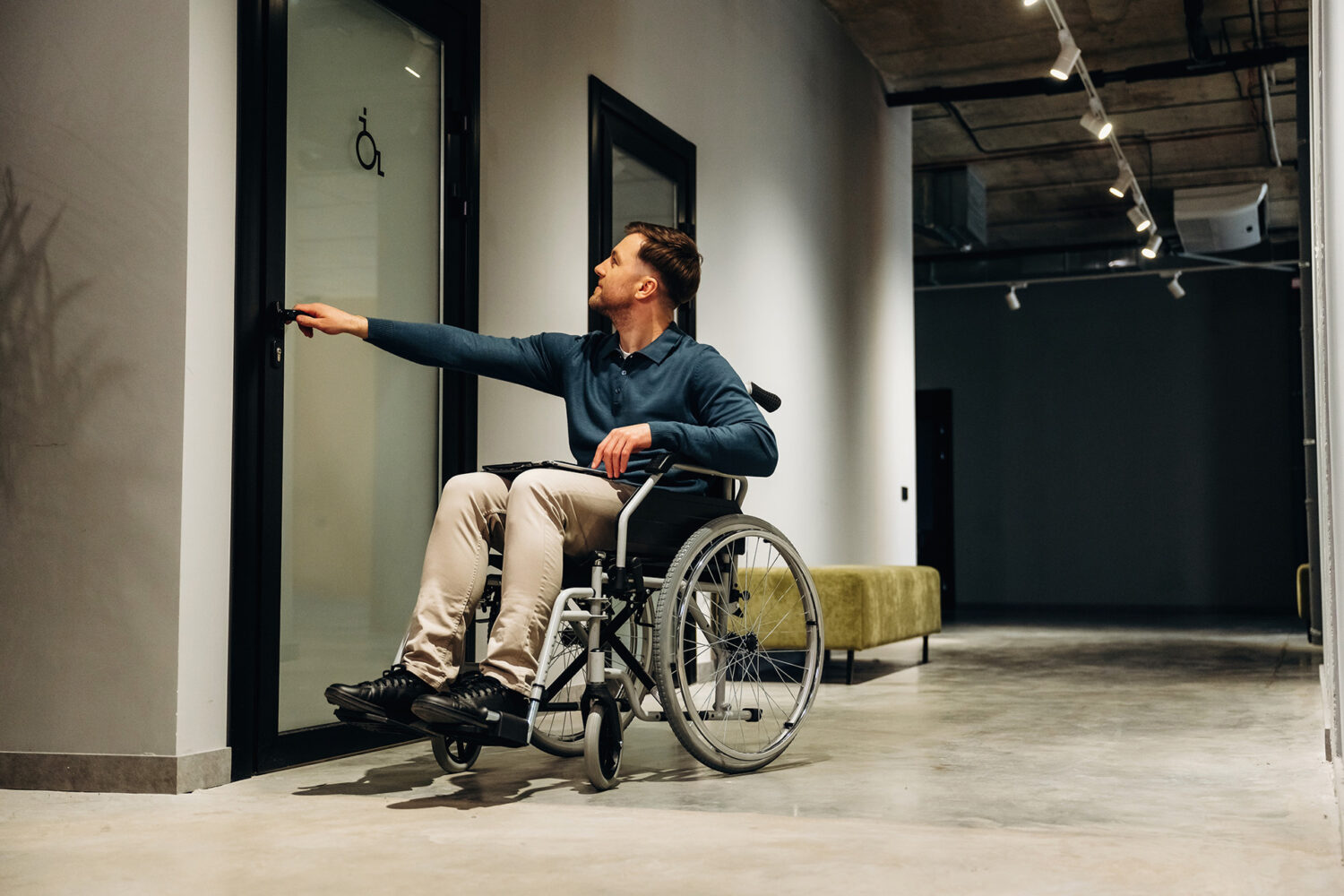 A man in a wheelchair reaches to open a door with an accessible restroom sign in a modern hallway with concrete floors and minimalist decor.