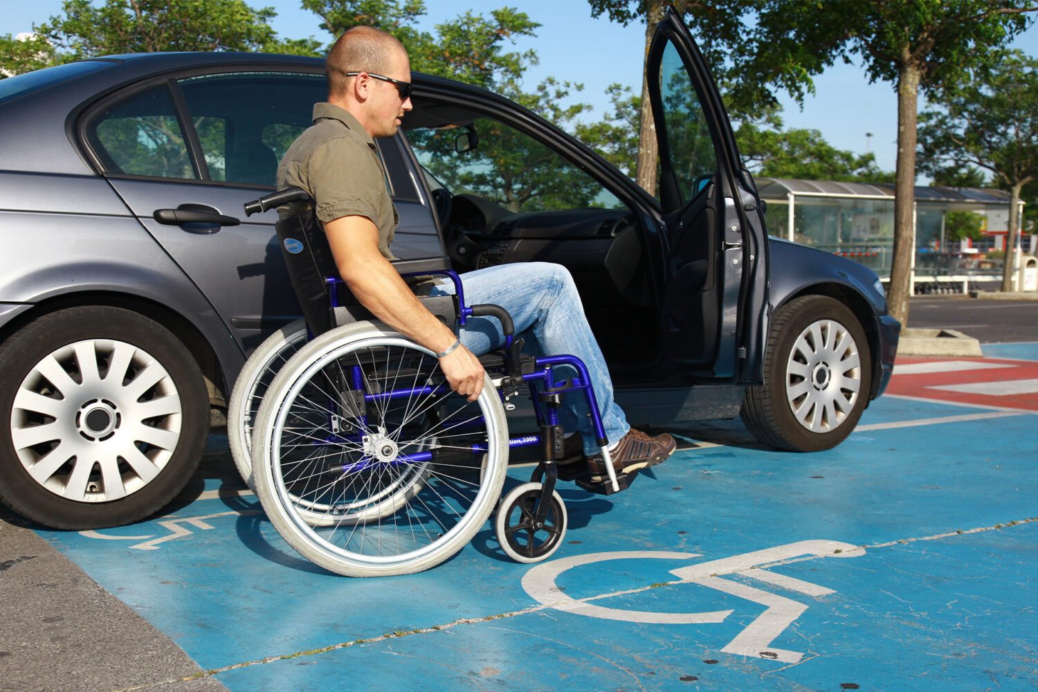 A man in a wheelchair is beside an open car door, positioned in a parking spot marked with a blue accessibility symbol.
