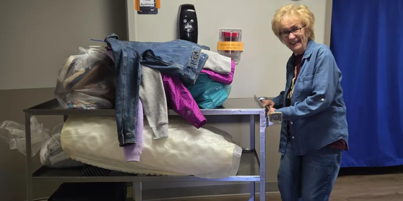 An older woman in glasses and a denim outfit smiles while standing next to a metal cart piled with bags and clothes, including jackets. The scene appears to be indoors, with medical equipment and a blue curtain in the background.