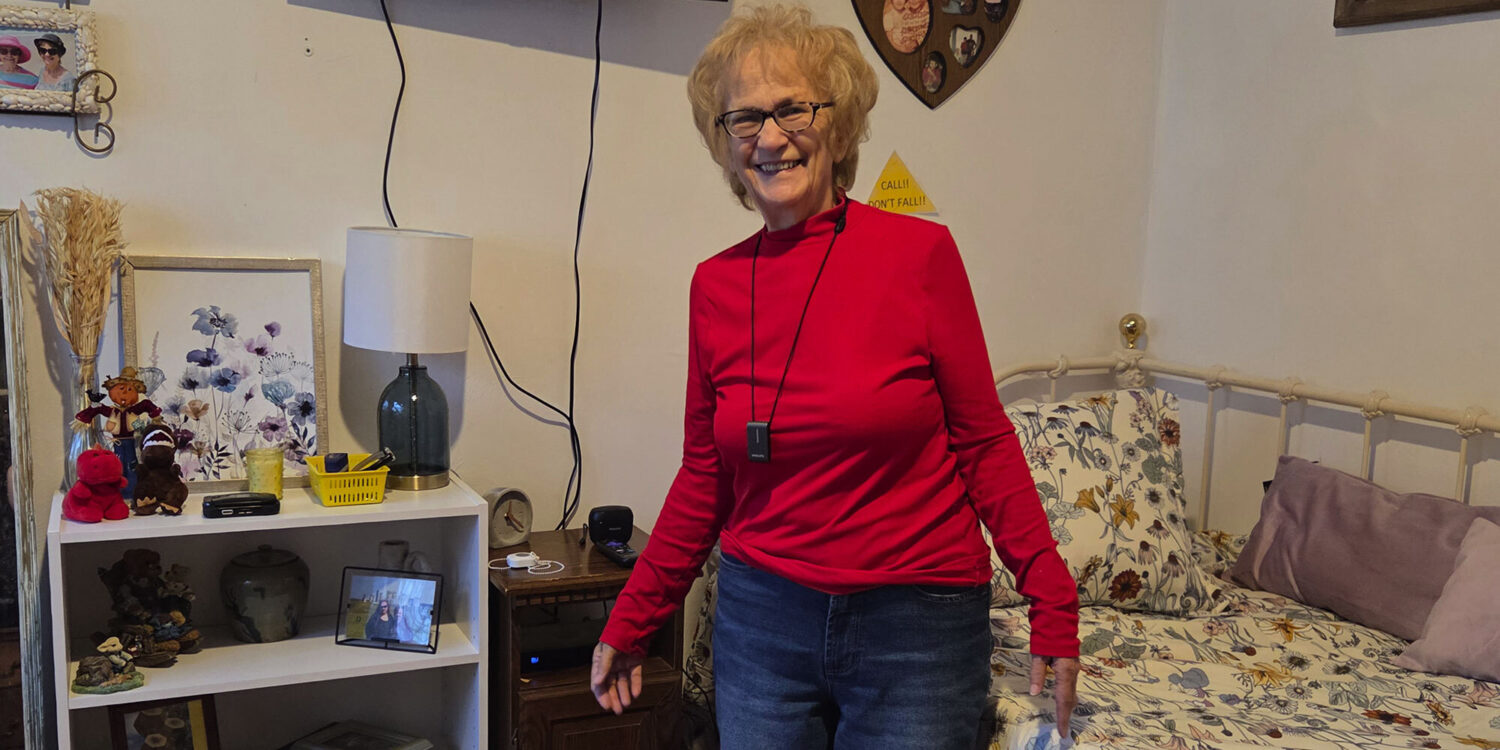 An elderly woman with glasses and a red long-sleeve shirt smiles while standing in a cozy bedroom with floral bedding, a white shelf with decor, and family photos on the wall.