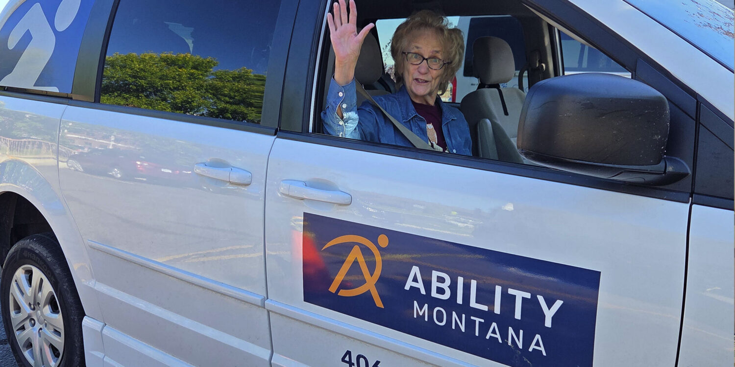 An older woman with glasses waves from the driver’s seat of a white van labeled “Ability Montana.” The door is open, and trees are reflected in the window.