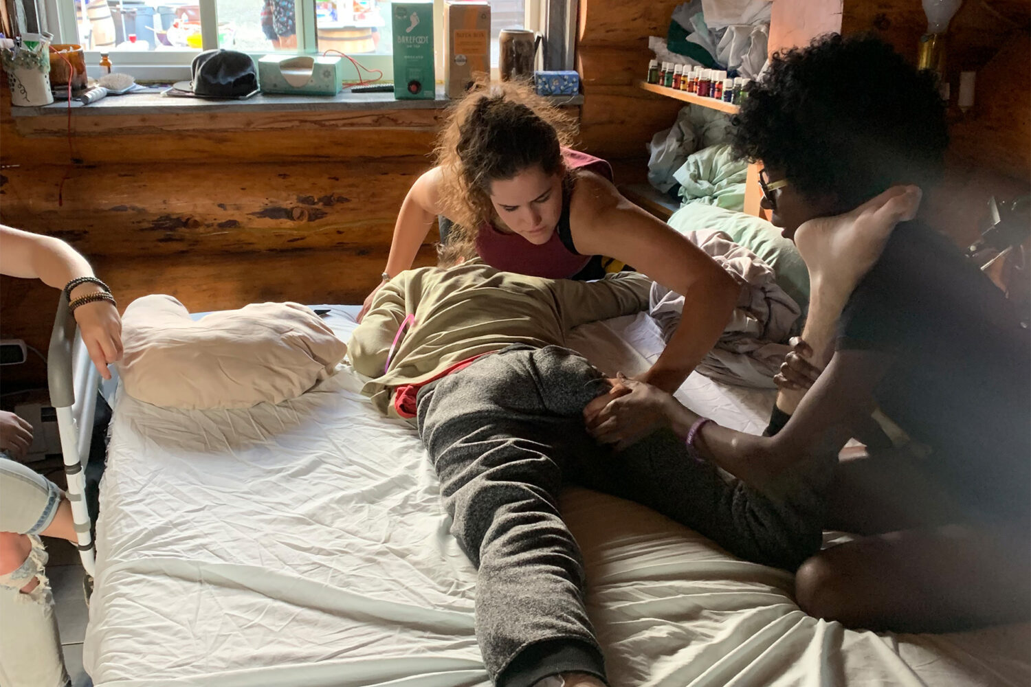 Two people on a bed assist a third person lying face down by stretching their leg and massaging their back, in a sunlit, wooden room with shelves and a window in the background.