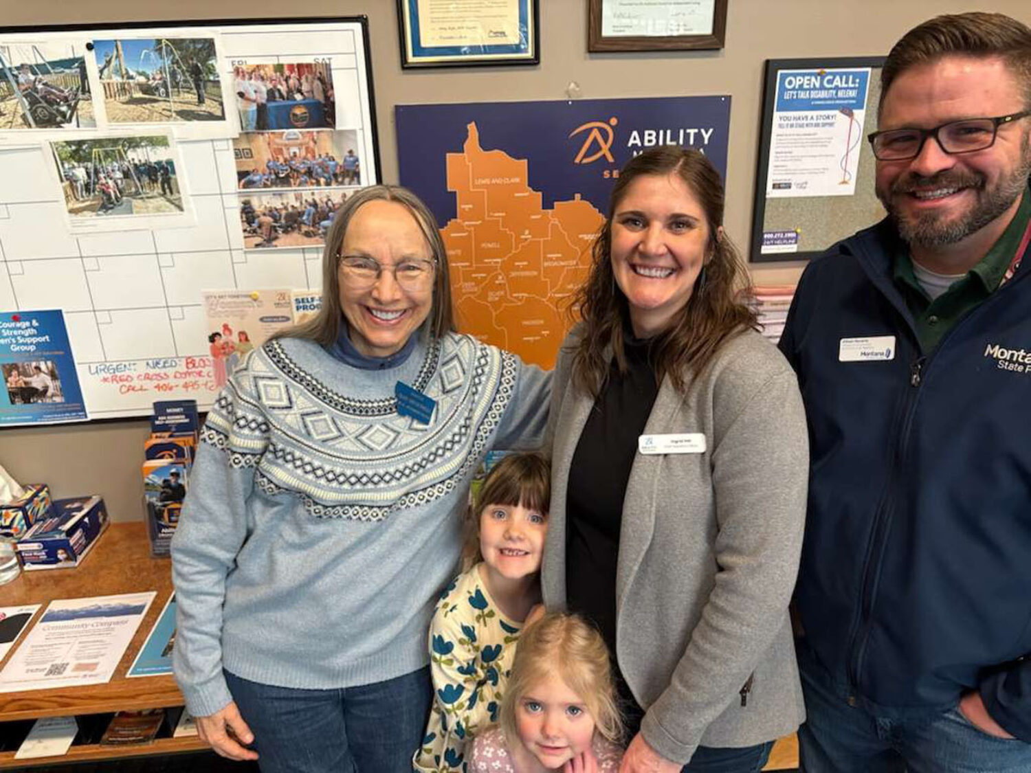 Four adults and two young girls smile together inside an office. Behind them are a wall calendar, informational posters, and a large orange map. The setting appears friendly and welcoming.