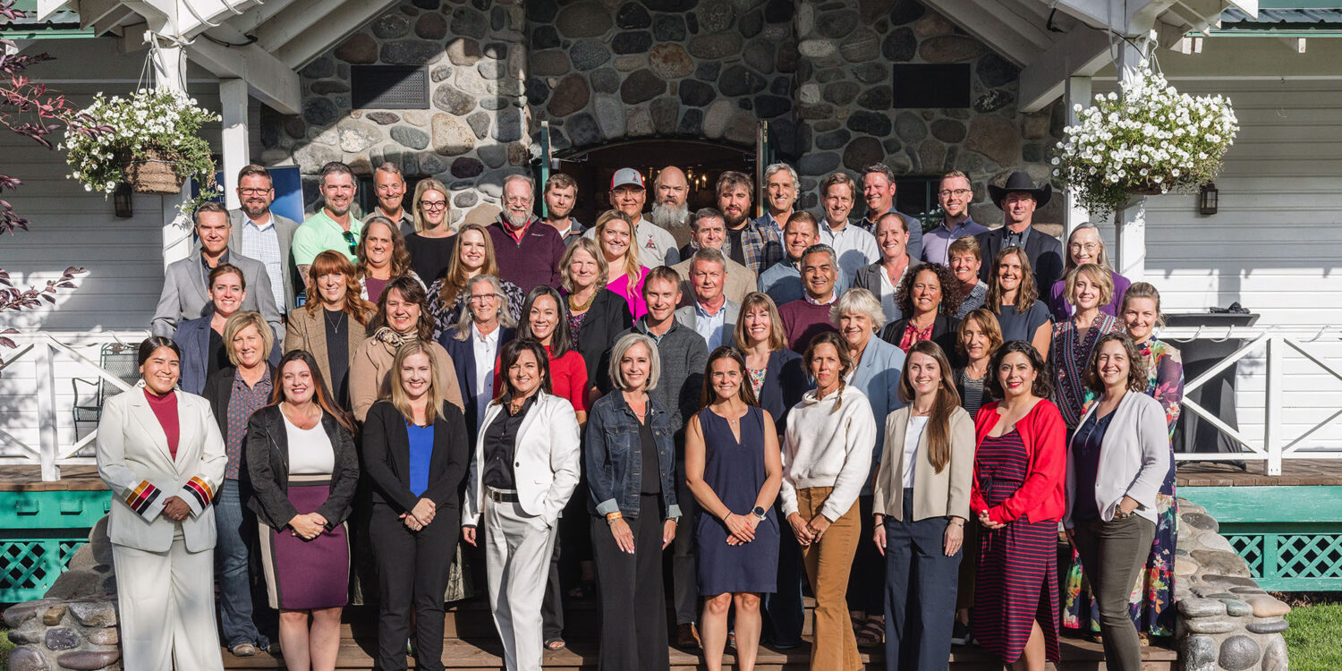 A large group of people, dressed in business and casual attire, posing together and smiling outside in front of a stone and white wood building with flowers hanging from the porch.