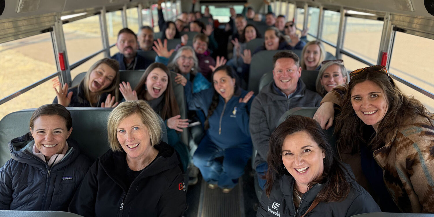 A group of smiling adults sit together on a school bus. Many are waving, laughing, or making playful gestures, creating a cheerful and lively atmosphere. The bus seats are visible, and the group appears excited and happy.