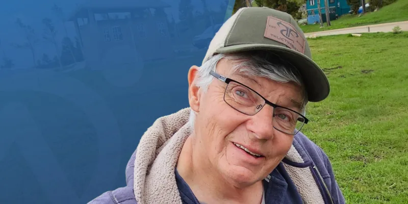 An older person wearing glasses, a green cap, and a jacket smiles outdoors on a grassy area, with trees and wooden cabins in the background and a blue graphic overlay on the left side.