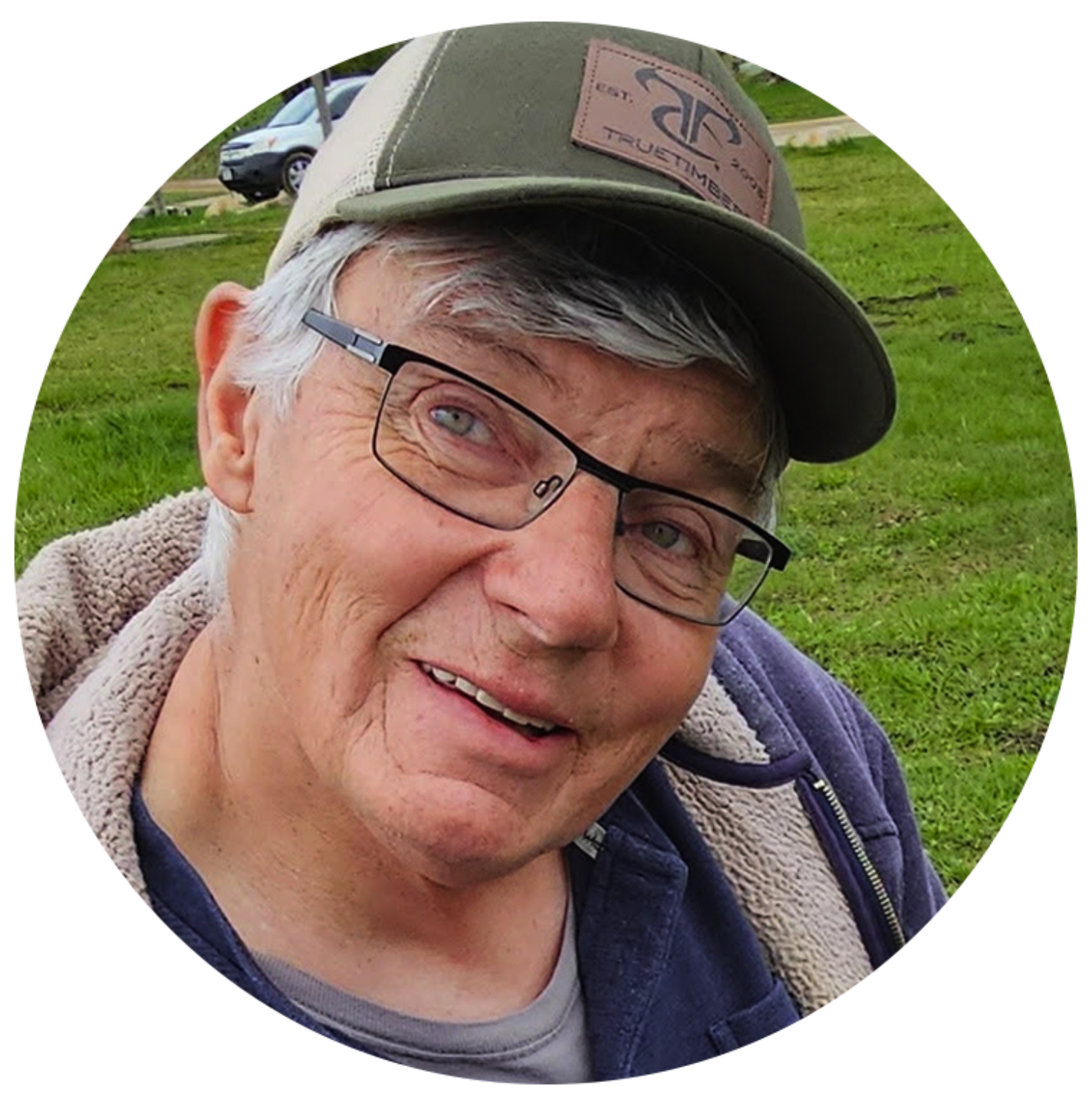 An older man with gray hair, glasses, and a green cap smiles warmly at the camera. He is outdoors, wearing a fleece jacket over a t-shirt, with green grass and a parked car in the background.
