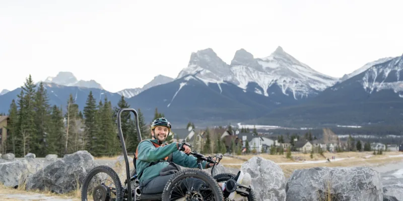 A smiling person in a green jacket sits in an adaptive mountain bike on a trail, with large rocks, trees, and snow-capped mountains in the background.