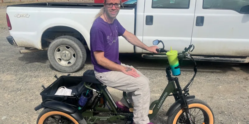 A person with long hair, glasses, and a purple shirt sits on a green electric tricycle with wide tires. The trike has a basket and drink holder. A white pickup truck and a barn are in the background.