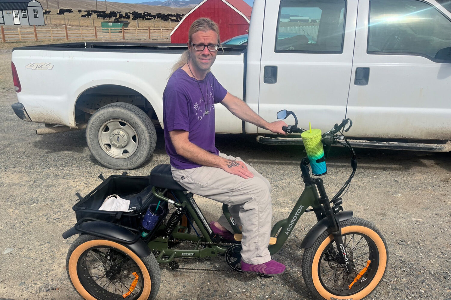A person with long hair, glasses, and a purple shirt sits on a green electric tricycle with wide tires. The trike has a basket and drink holder. A white pickup truck and a barn are in the background.