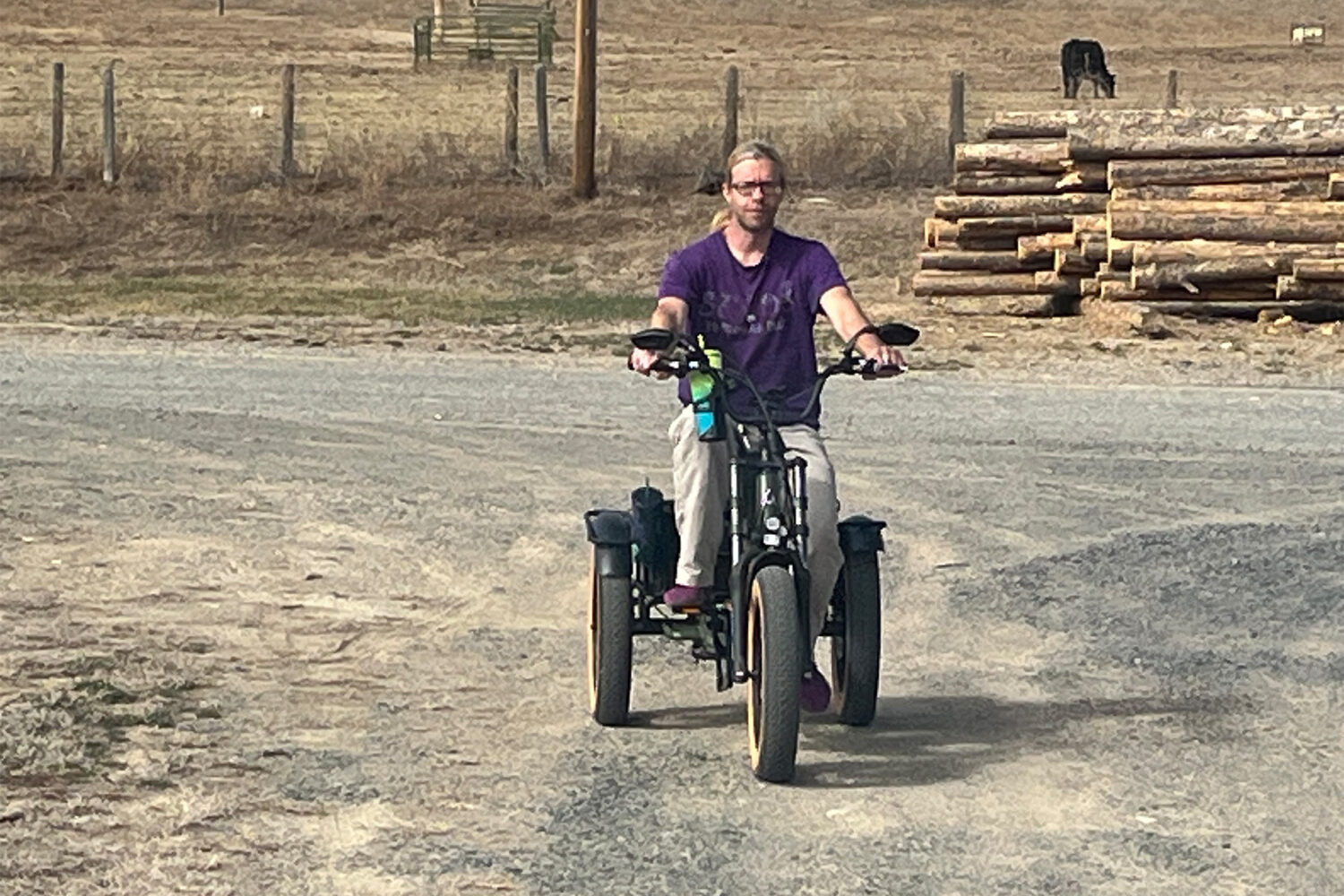 A person with long hair and glasses rides an electric tricycle on a dirt path in a rural area. Stacks of logs, fencing, and grazing cows are visible in the background.