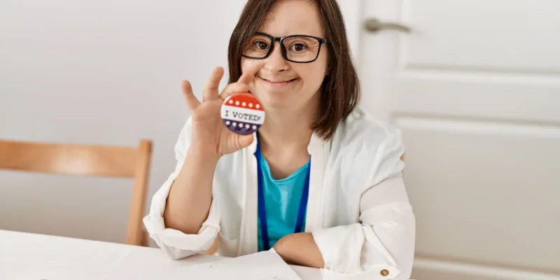 A woman with Down syndrome smiles and holds up an "I Voted" sticker while sitting at a table with a clipboard, a paper, a pen, and a phone. She is wearing glasses and a white jacket.