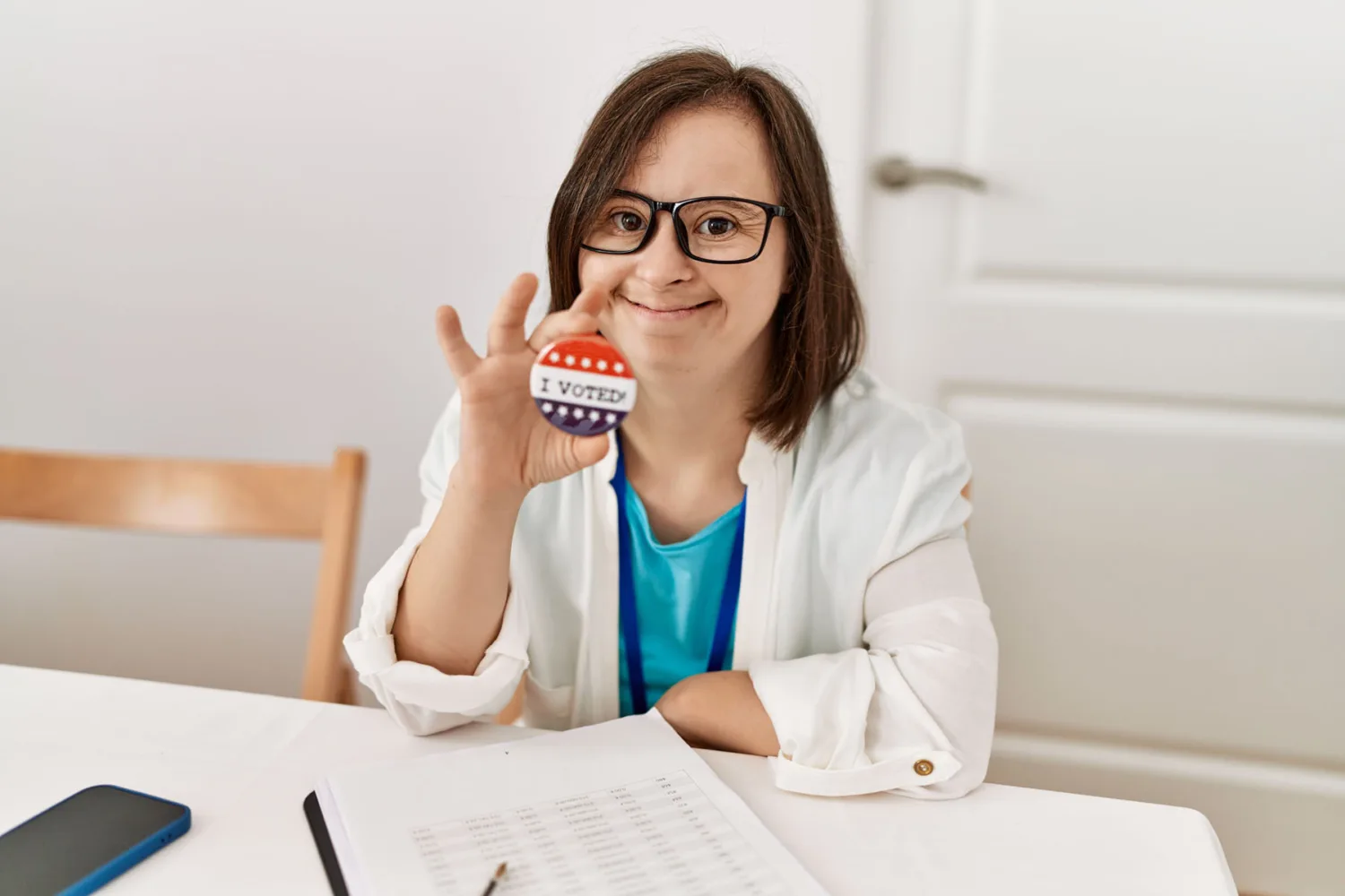 A woman with Down syndrome smiles and holds up an "I Voted" sticker while sitting at a table with a clipboard, a paper, a pen, and a phone. She is wearing glasses and a white jacket.
