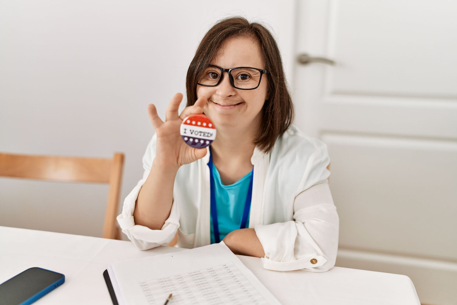 A woman with Down syndrome smiles and holds up an "I Voted" sticker while sitting at a table with a clipboard, a paper, a pen, and a phone. She is wearing glasses and a white jacket.