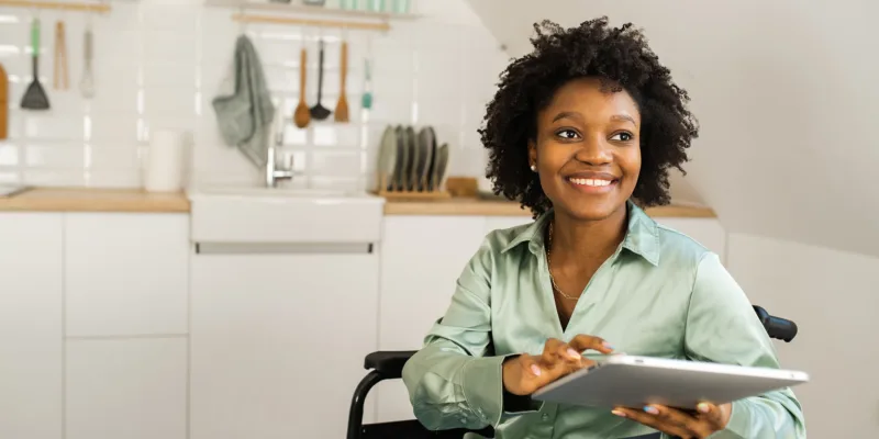 A smiling woman sitting in a wheelchair uses a tablet in a bright, modern kitchen with shelves, dishes, and utensils visible in the background.