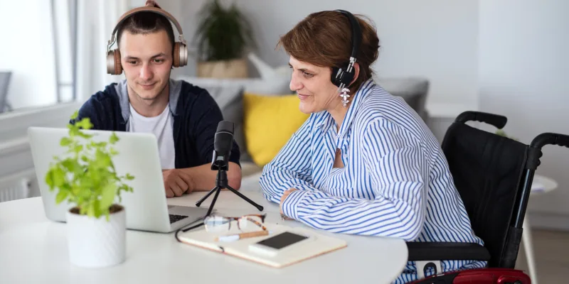 A young man and a woman in a wheelchair, both wearing headphones, sit at a table with a laptop, microphone, notebook, and phone, recording or participating in a podcast in a bright room.