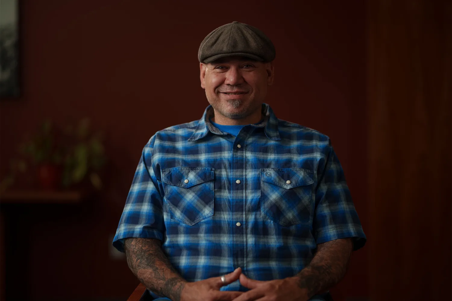 Charles Alvarez of Ability Montana sits smiling, with hands clasped. A plant is visible in the dimly lit background.