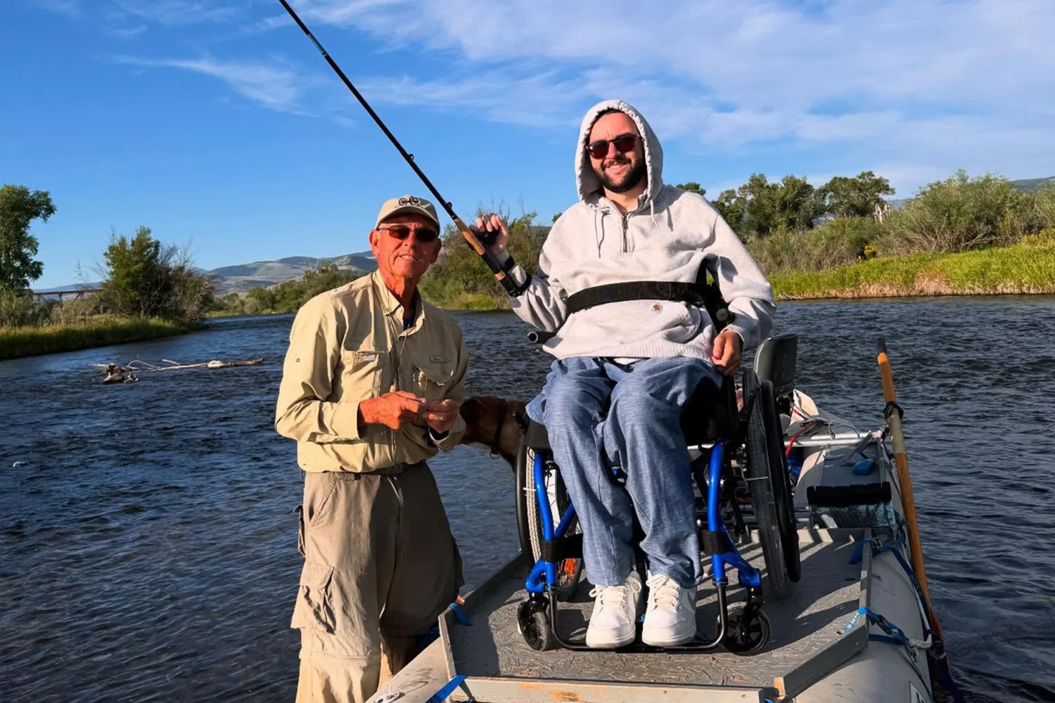 Two men on a boat fishing in a river; one is standing and wearing sunglasses and a hat, the other is smiling in a wheelchair holding a fishing rod. Trees and mountains are visible in the background.