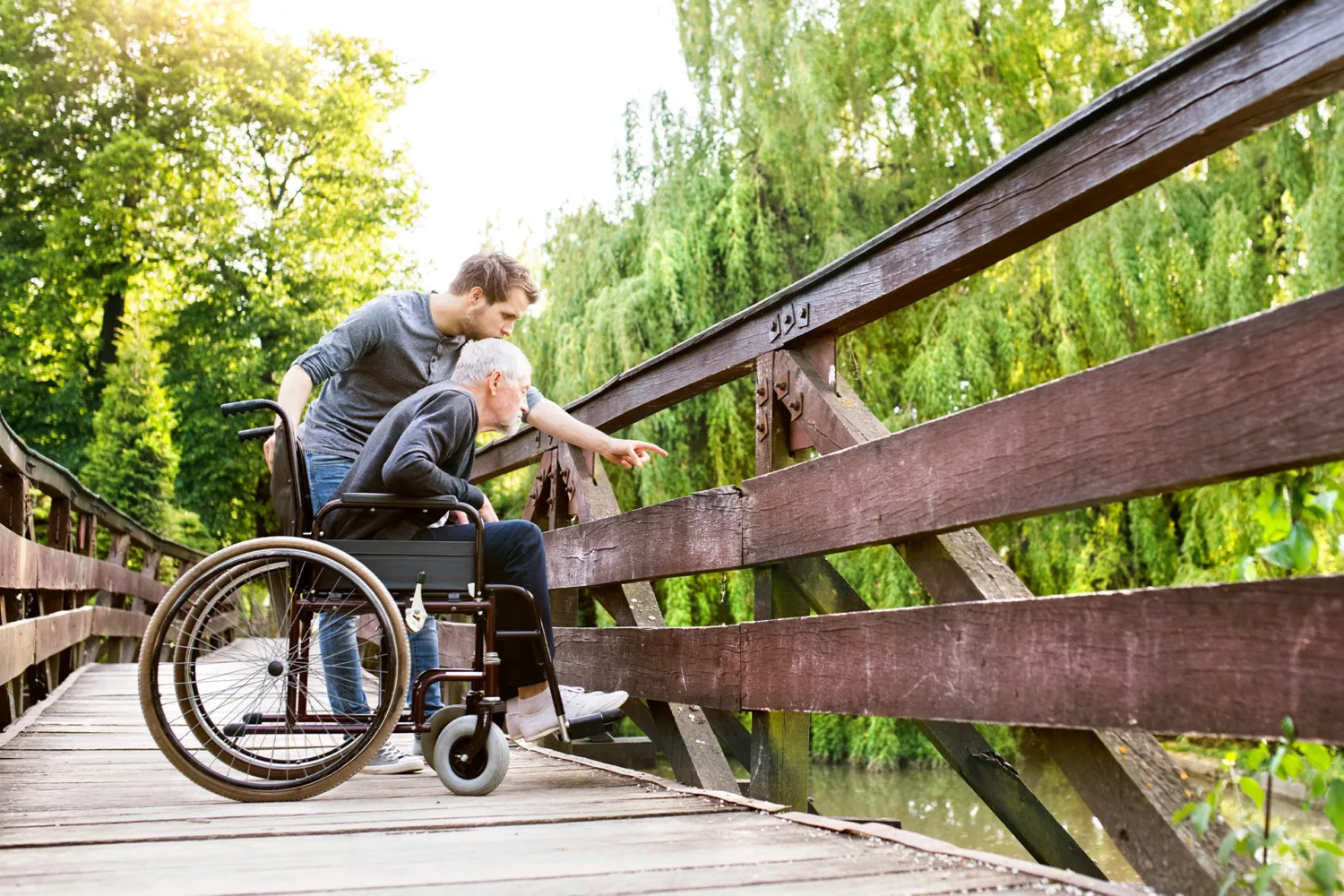 A young man stands behind an older man in a wheelchair on a wooden bridge, both looking and pointing toward something below, surrounded by lush green trees.