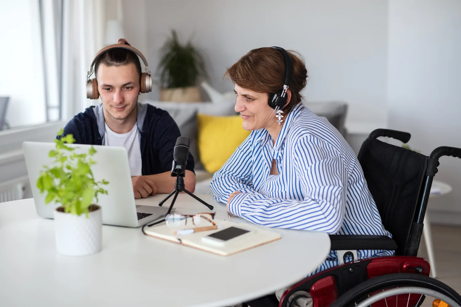 A young man and a woman in a wheelchair, both wearing headphones, sit at a table with a laptop, microphone, notebook, and phone, recording or participating in a podcast in a bright room.