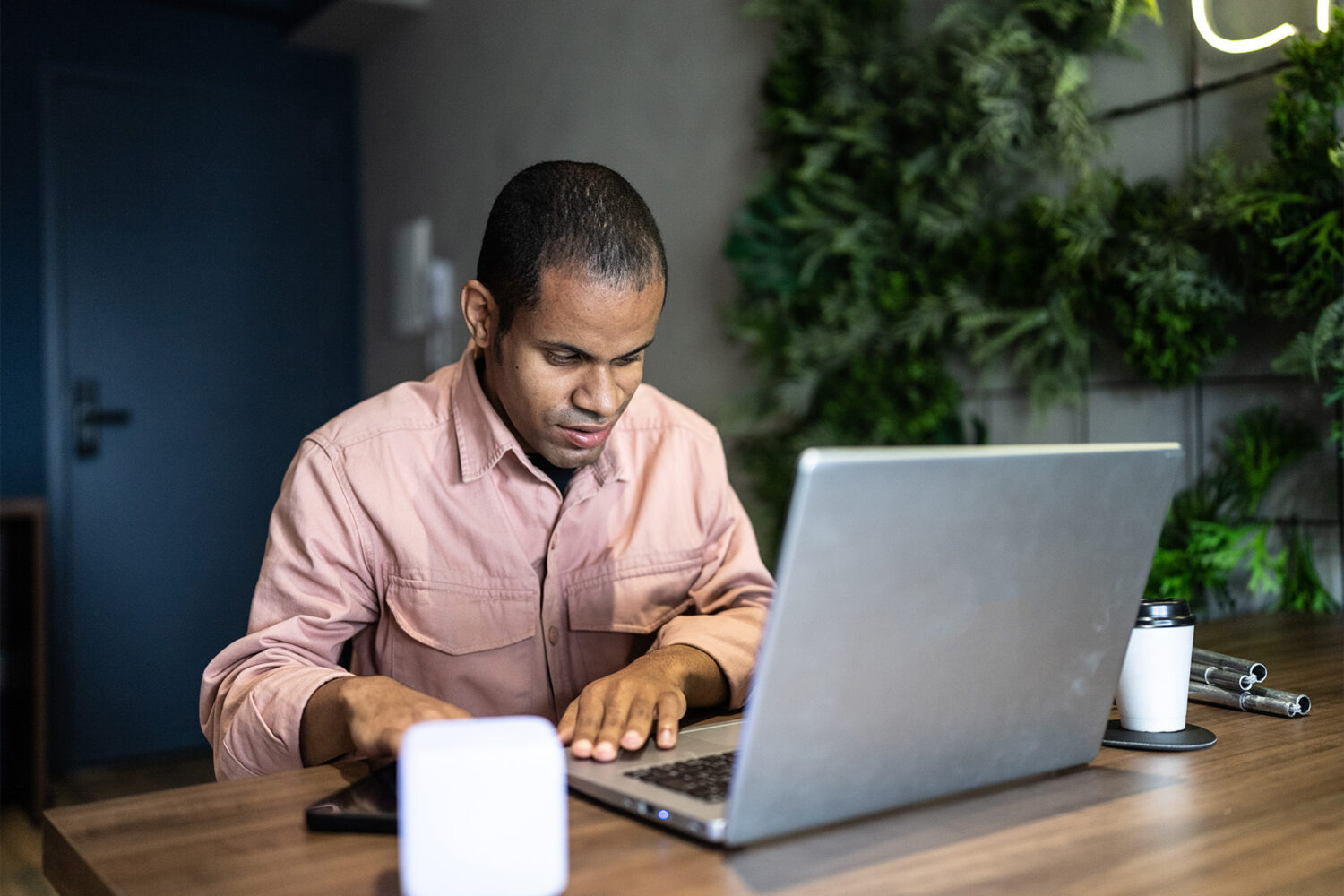 A man, sitting at a desk with a laptop and tablet, works intently. There is a cup of coffee and a green wall with plants in the background.