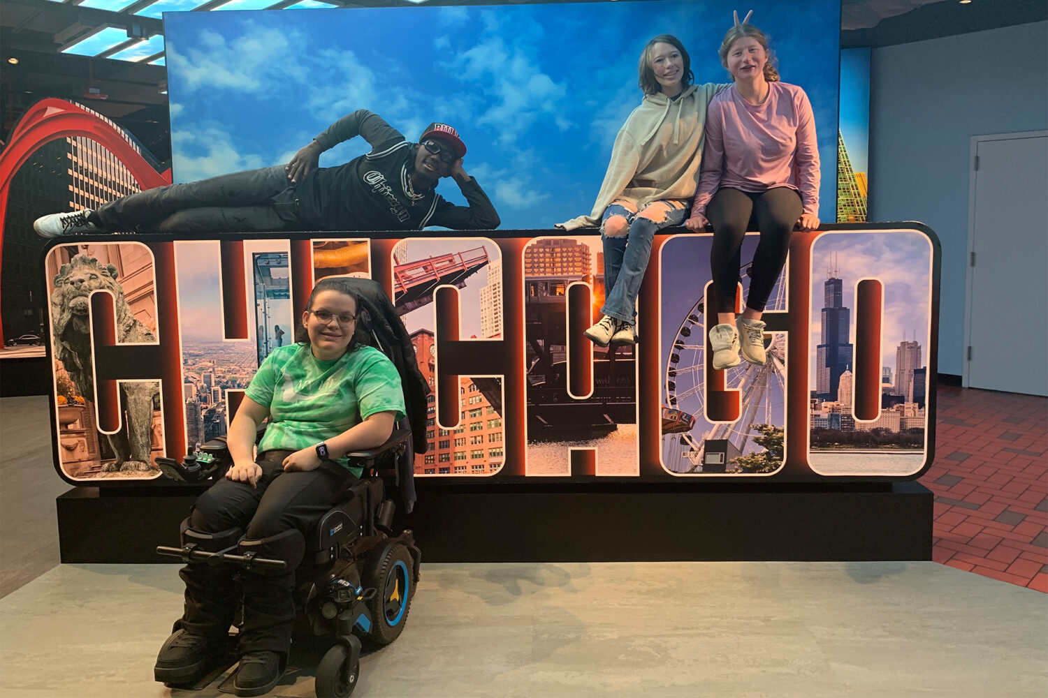 Four people pose around a large "Chicago" sign; one lies on top, two sit on it, and one in a wheelchair is in front. The background depicts city buildings.