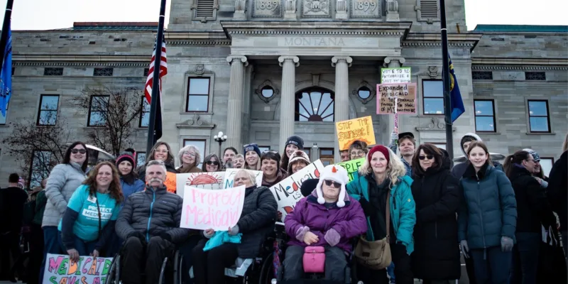 A diverse group of people, some in wheelchairs, hold colorful protest signs advocating for Medicaid in front of a government building with columns and “Montana” inscribed above the entrance.