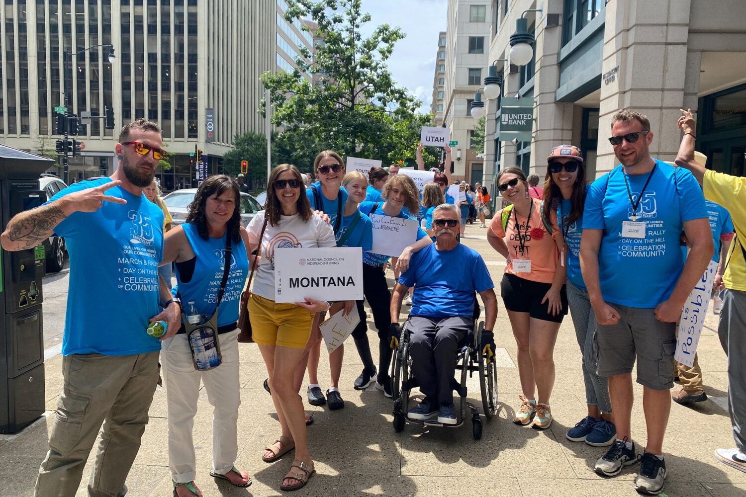 A group of people stand smiling on a city sidewalk, some wearing blue shirts and sunglasses. One person in a wheelchair holds a "Montana" sign, indicating the group represents Montana at an event.
