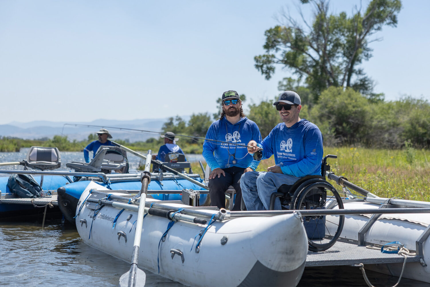 Two men in blue shirts, one in a wheelchair, fish from an inflatable boat on a river. Other people in similar boats are nearby. The scene is outdoors with green grass, trees, and mountains in the background.