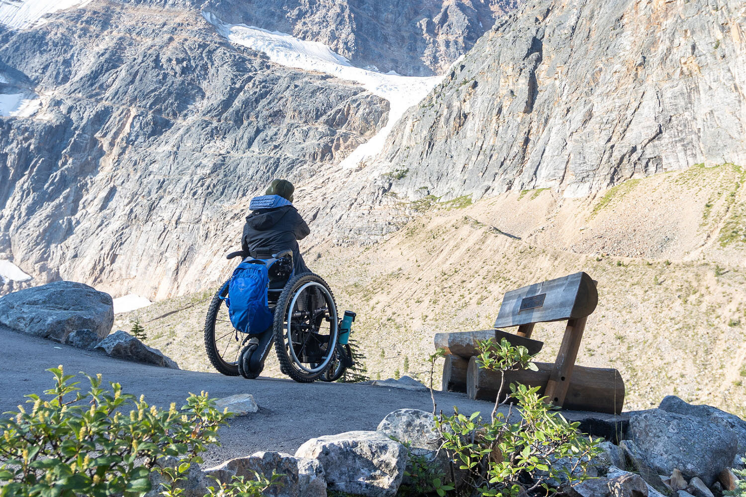 A person in a wheelchair looks at a mountain landscape next to a wooden bench on a paved path. Rocks, shrubs, and steep cliffs are visible under bright sunlight.