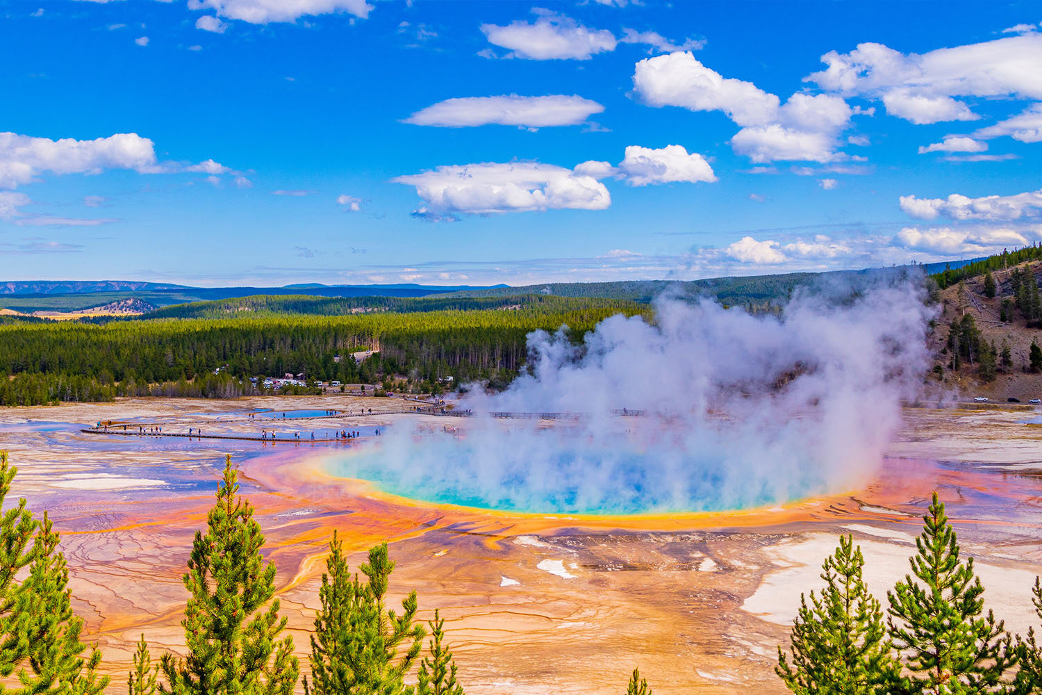 Aerial view of Grand Prismatic Spring in Yellowstone National Park, showing vibrant blue, yellow, and orange colors surrounded by steam and green forest under a partly cloudy sky.