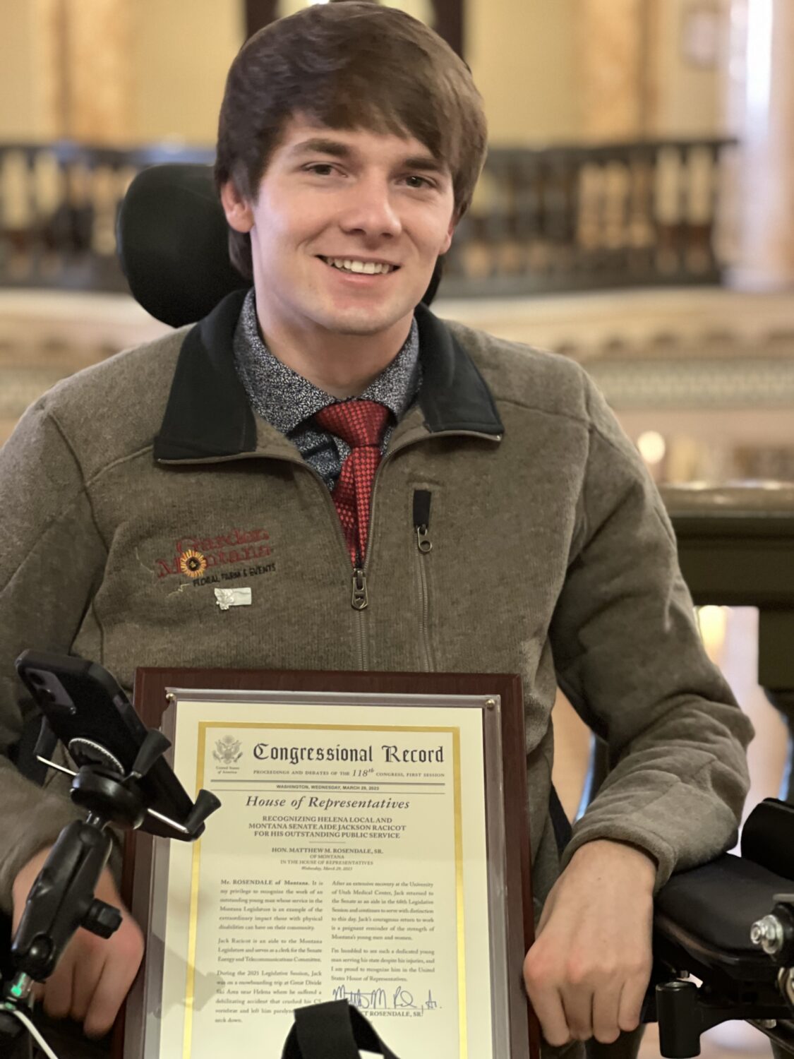 A young man is sitting in a motorized wheelchair, holding a framed Congressional Record certificate. He is indoors, with ornate architectural details visible in the background.