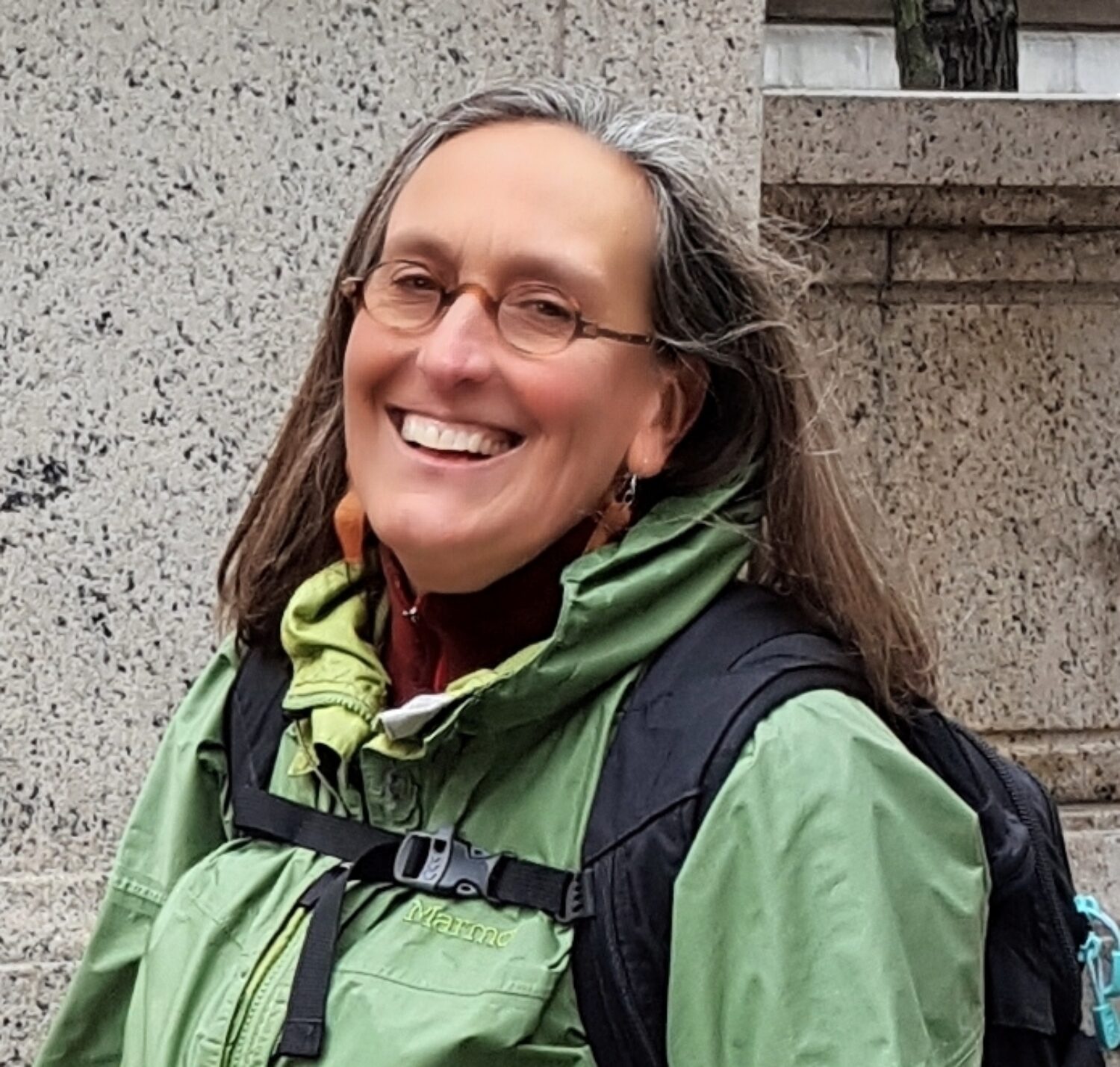 A woman with long hair wearing glasses, a green jacket, and a backpack smiles in front of a stone wall.