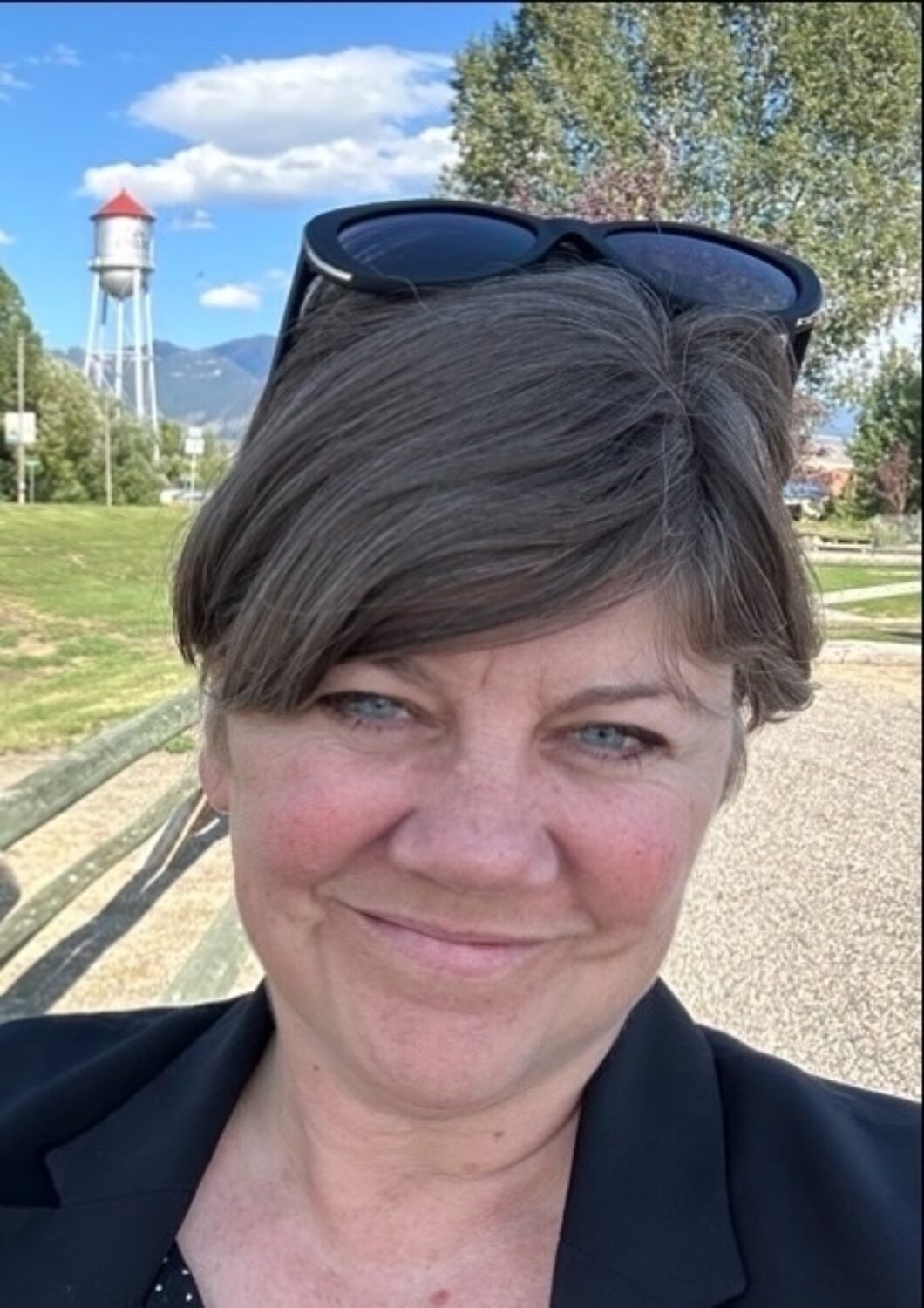 A person with short hair and sunglasses atop their head smiles at the camera, with a water tower and trees in the background under a partly cloudy sky.