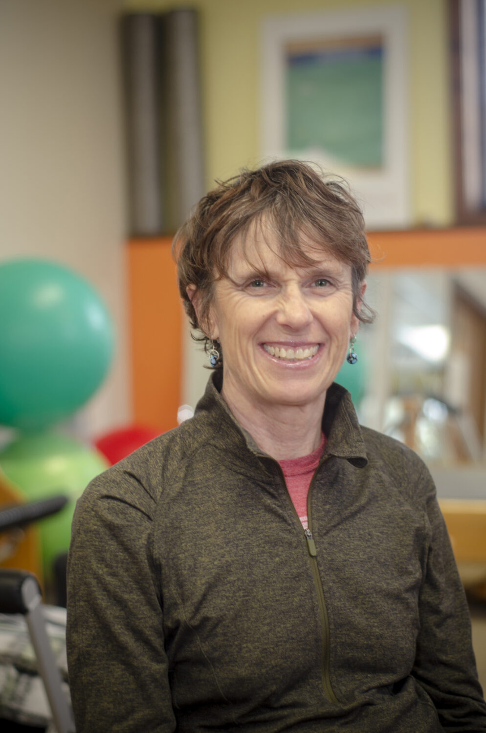 A middle-aged person with short hair smiles while sitting indoors, wearing a zipped-up jacket. A green exercise ball and mirror are in the background.