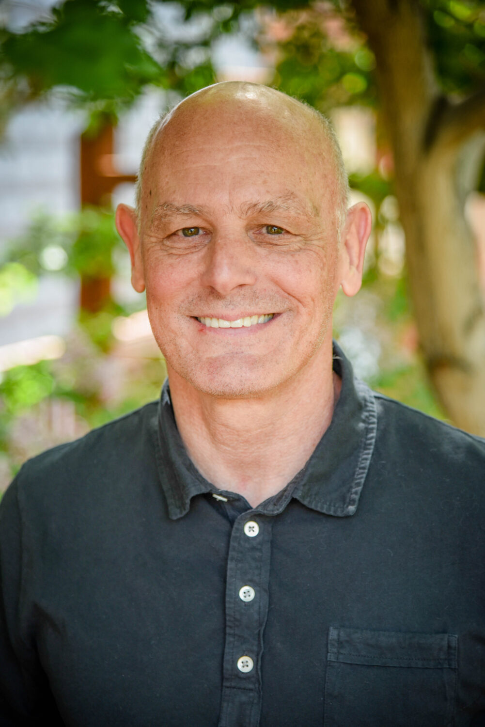 A bald man wearing a dark collared shirt smiles while standing outdoors with greenery in the background.