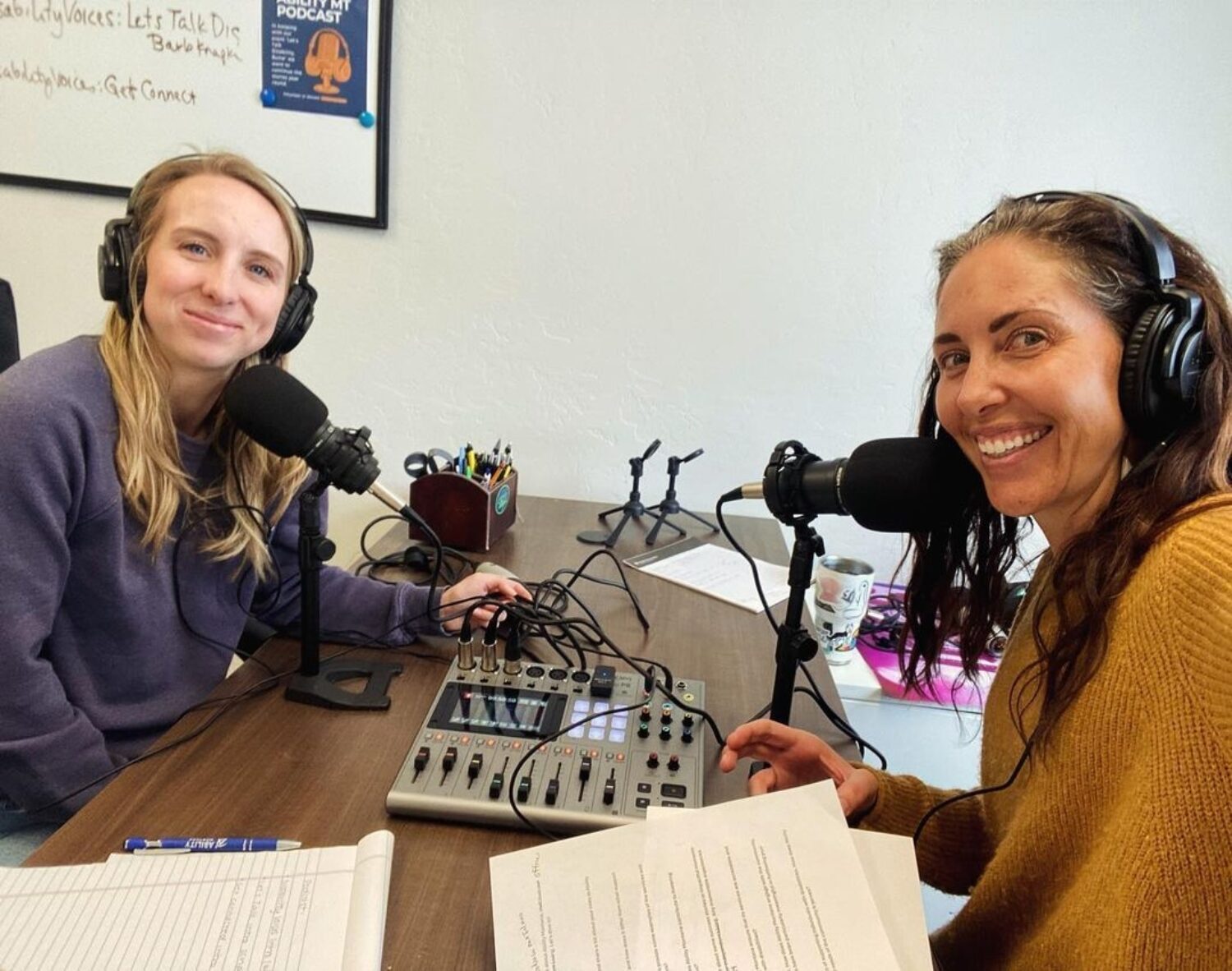 Two women smiling while recording a podcast with microphones, headphones, and audio equipment on a table.