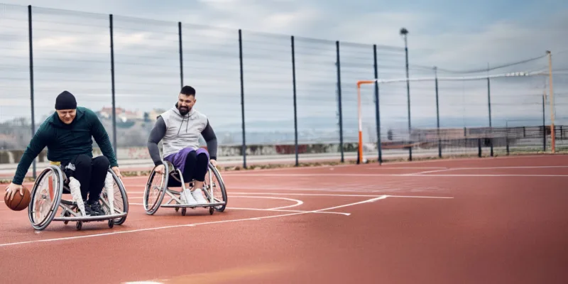 Two men in wheelchairs play basketball on an outdoor court. One man dribbles the ball while the other smiles, both enjoying the game. The court is enclosed by a tall fence and the sky is overcast.