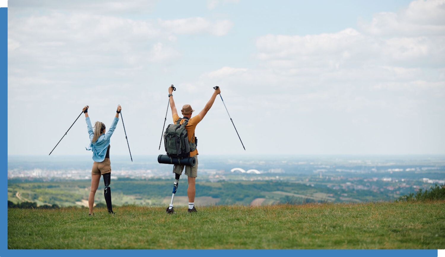 Two hikers with prosthetic legs stand on a grassy hill, raising their trekking poles in celebration while facing a scenic landscape with a city and hills in the distance under a partly cloudy sky.