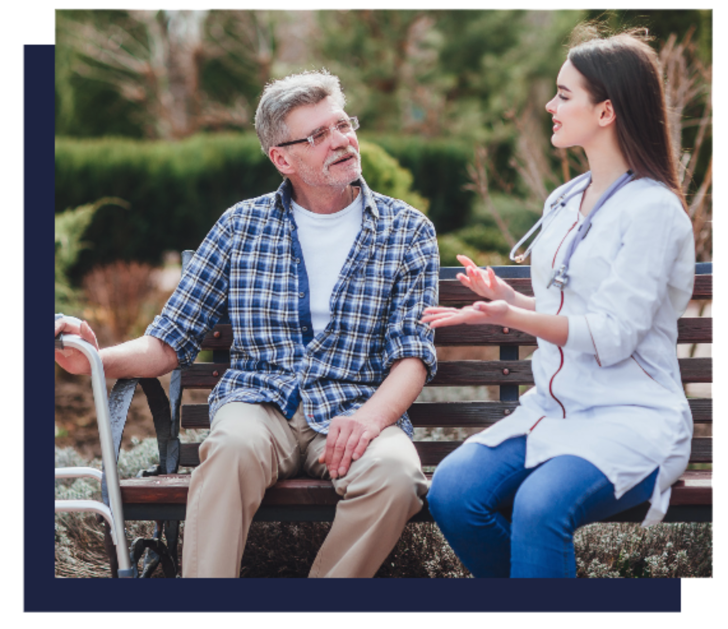An older man with a cane sits on a bench outdoors, attentively listening to a young female healthcare worker who is speaking and gesturing with her hands.