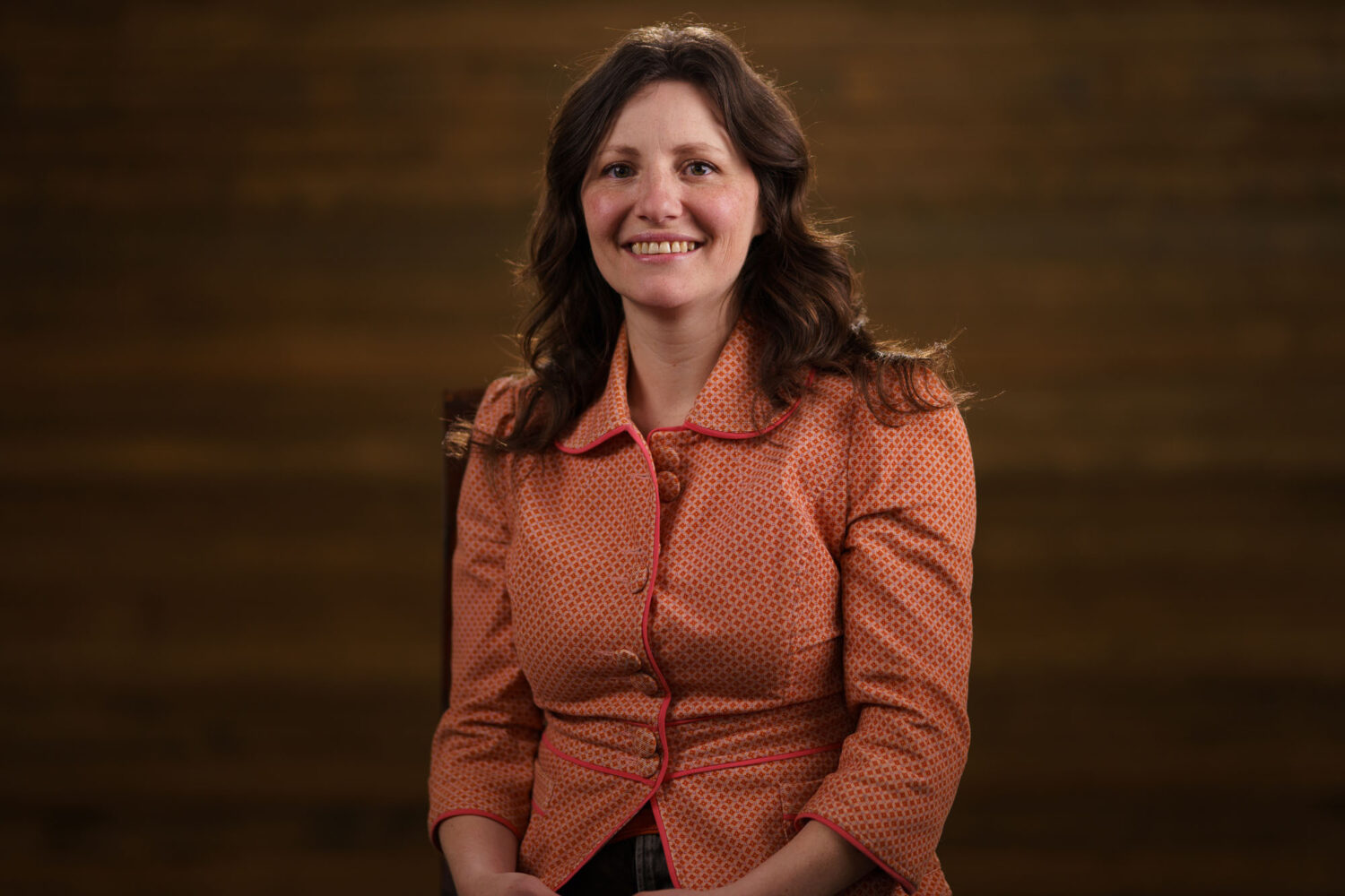 A woman in an orange jacket smiling in front of a wooden background. profile picture