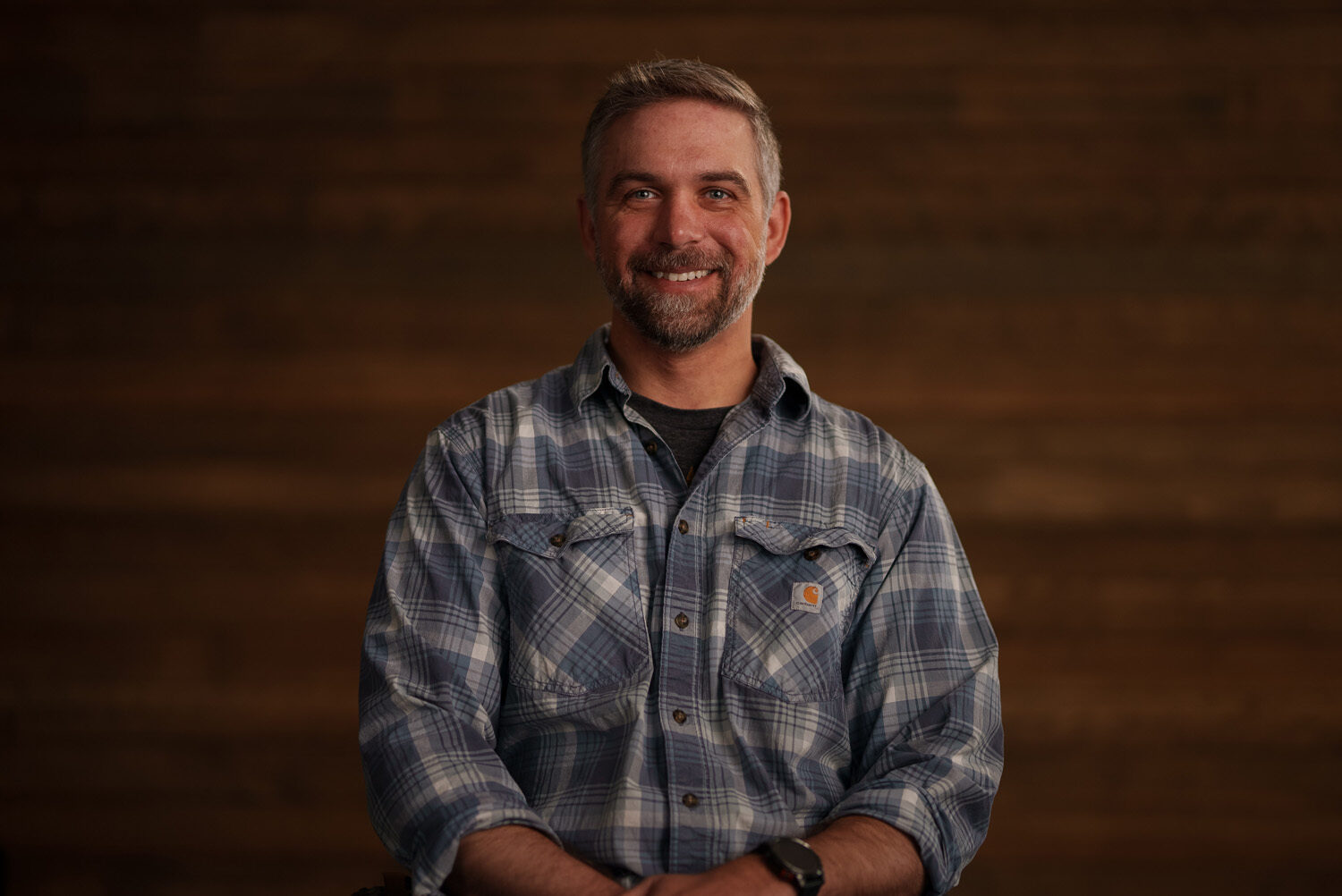A man in a plaid shirt smiling in front of a wooden wall. profile picture