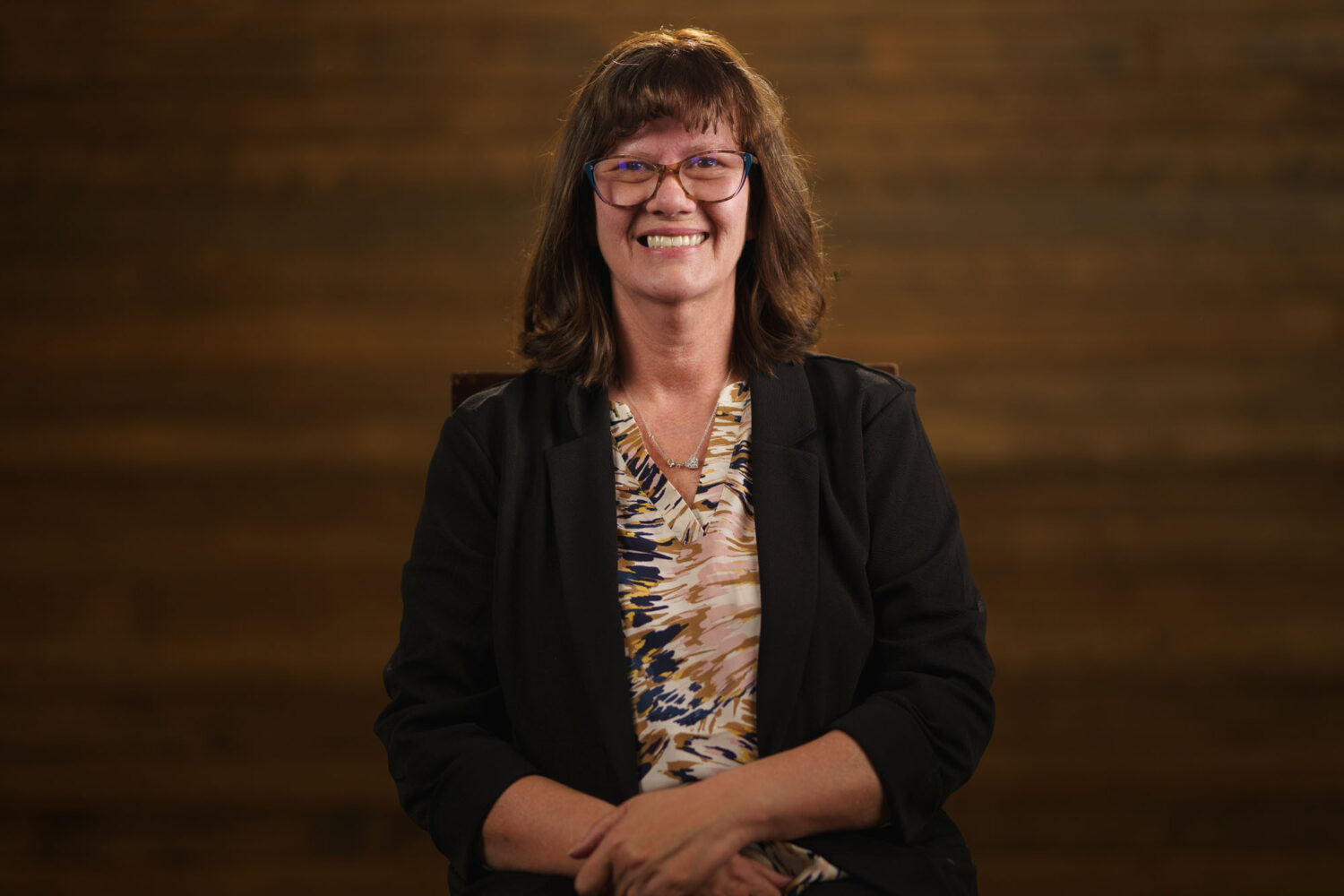 A woman with glasses smiling in front of a wooden background. profile picture