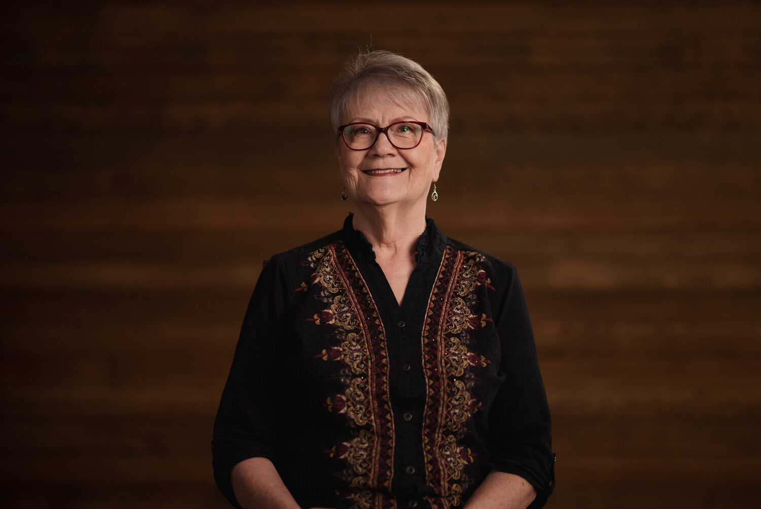 A woman in glasses is smiling in front of a wooden background. profile picture