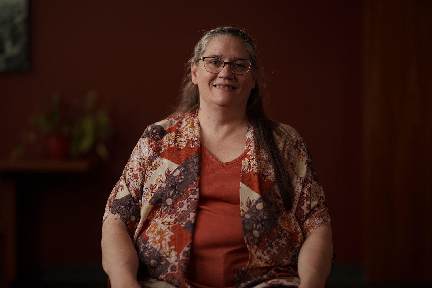 A woman wearing glasses and a floral shirt sitting in front of a red wall. profile picture